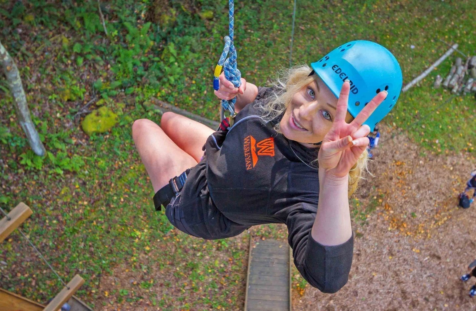A college student flashes a peace sign while suspended in the air over the Team Challenge Course at ACE Adventure Resort