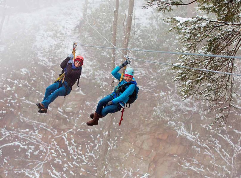 Two women fly through the trees of the New River Gorge winter at ACE Adventure Resort. Holiday vacations are the best!